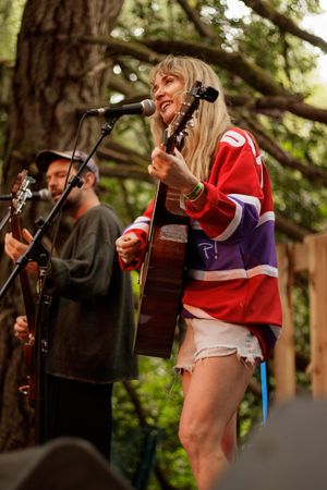 Blonde woman in red jersey plays acoustic guitar and sings outdoors at a music festival with trees in background.