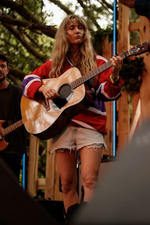 Female musician in red jersey playing acoustic guitar on outdoor stage in shorts.