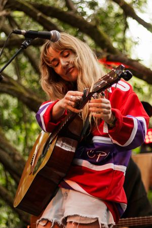 Woman in red hockey jersey playing acoustic guitar at outdoor music festival.