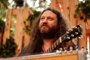 Long-haired bearded musician playing acoustic guitar outdoors, looking upward thoughtfully near wooden backdrop.