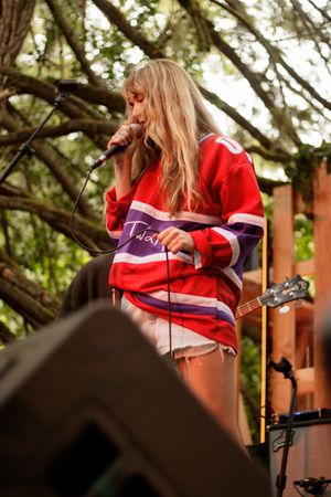 Blonde female singer in red hockey jersey performing with microphone on outdoor stage.