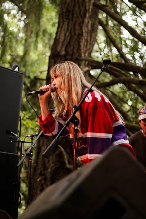 Female vocalist singing into microphone on outdoor stage surrounded by trees.