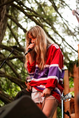 Blonde female singer performing on outdoor stage wearing red and purple hockey jersey with microphone.