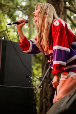 Blonde female singer performing on outdoor stage wearing red hockey jersey, singing passionately into microphone at music festival.