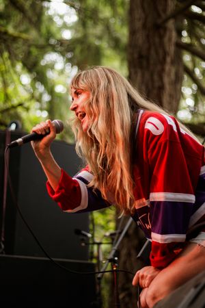Blonde female singer performing outdoors at festival, leaning into microphone wearing red hockey jersey with trees in background.