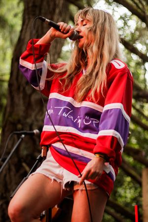Female singer performing on outdoor stage wearing red and purple Shania Twain jersey with denim shorts, singing into microphone.