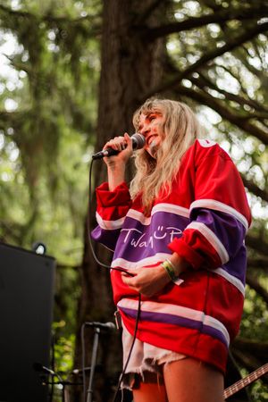 Blonde female singer performing on outdoor stage wearing red purple hockey jersey holding microphone among trees.