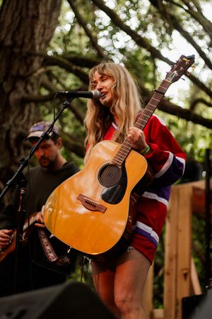 Blonde female musician playing acoustic guitar and singing into microphone on outdoor stage surrounded by trees.
