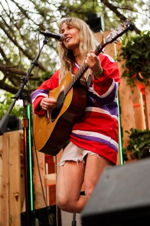 Smiling blonde woman playing acoustic guitar on outdoor stage wearing red hockey jersey and denim shorts.