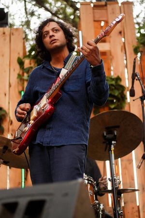 Male guitarist playing red electric guitar on outdoor stage with wooden backdrop and cymbal visible.