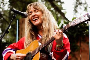 Joyful blonde woman strumming acoustic guitar outdoors, laughing while performing in a red jersey.