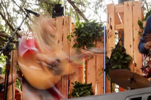 Blond musician playing acoustic guitar on outdoor stage with wooden backdrop and green foliage decorations.