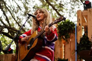 Blonde woman playing acoustic guitar and singing at outdoor music performance, wearing colorful striped sweater.
