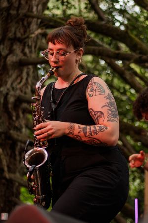 Tattooed woman playing saxophone outdoors at a music festival, wearing black sleeveless top, trees in background.