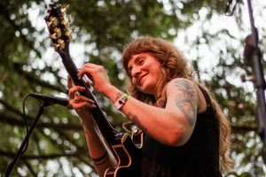 Smiling female guitarist with tattoos plays electric guitar outdoors at a live music festival.