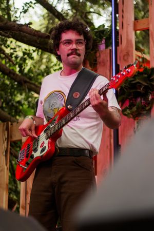 Man with mustache and glasses playing red electric bass guitar on outdoor stage.