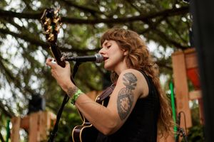 Woman with tattoos playing acoustic guitar and singing into microphone outdoors at music festival.