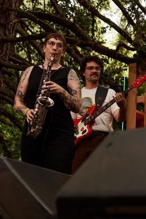 Tattooed woman playing saxophone on outdoor stage with male guitarist in background surrounded by trees.