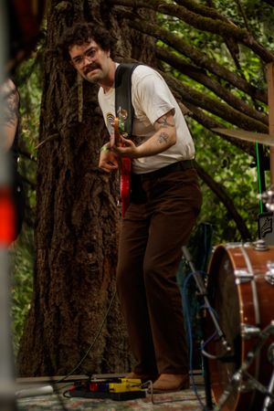 Bearded man with glasses plays red electric guitar leaning against large tree trunk at outdoor concert.