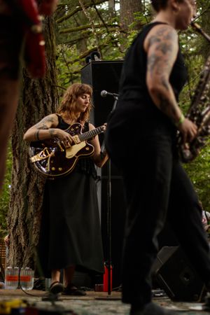Tattooed female guitarist in black dress performs outdoors at forest stage music festival.