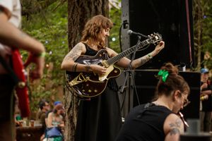 Tattooed woman playing black hollow-body guitar performing on outdoor forest stage.