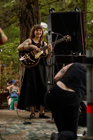 Woman in black dress playing acoustic guitar on outdoor stage surrounded by trees at a music festival.