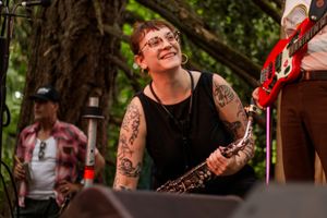 Tattooed woman playing saxophone outdoors at music festival, smiling joyfully with guitarist visible in background.