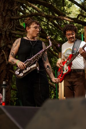 Tattooed woman playing saxophone on outdoor stage beside guitarist in wooded festival setting.