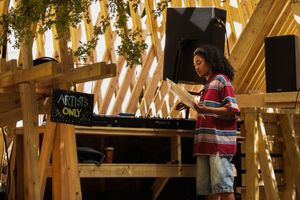Young DJ performing at an artists-only outdoor wooden stage venue with speakers and hanging greenery.