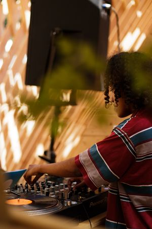 Young female DJ with curly hair mixing tracks on Pioneer turntables in warm-lit venue wearing striped shirt.