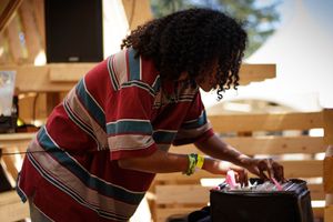 A person with curly hair in a striped red shirt leans over a black case outdoors near wooden pallets.