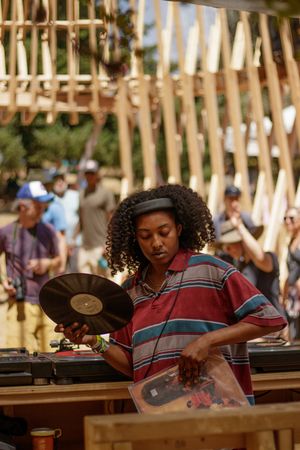A woman with curly hair holds a vinyl record at an outdoor DJ setup, wearing a striped polo shirt at a festival.