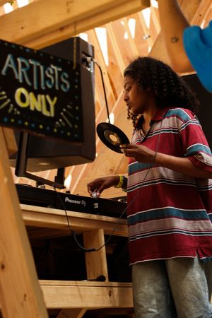 Young DJ holding vinyl record at Pioneer DJ turntable setup beneath an Artists Only neon sign.