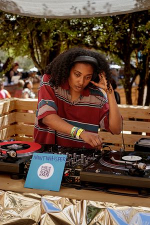 Female DJ mixing records at outdoor event, wearing striped shirt and headphones, with Mint Whisper sign visible.