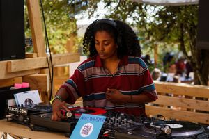 A female DJ mixing tracks on turntables at an outdoor music event, wearing a striped shirt.