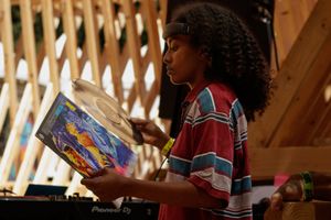Young DJ with curly hair holds colorful vinyl record at outdoor music festival near Pioneer DJ equipment.