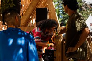 People building a wooden structure outdoors in sunlight, collaborating on a carpentry project surrounded by trees.