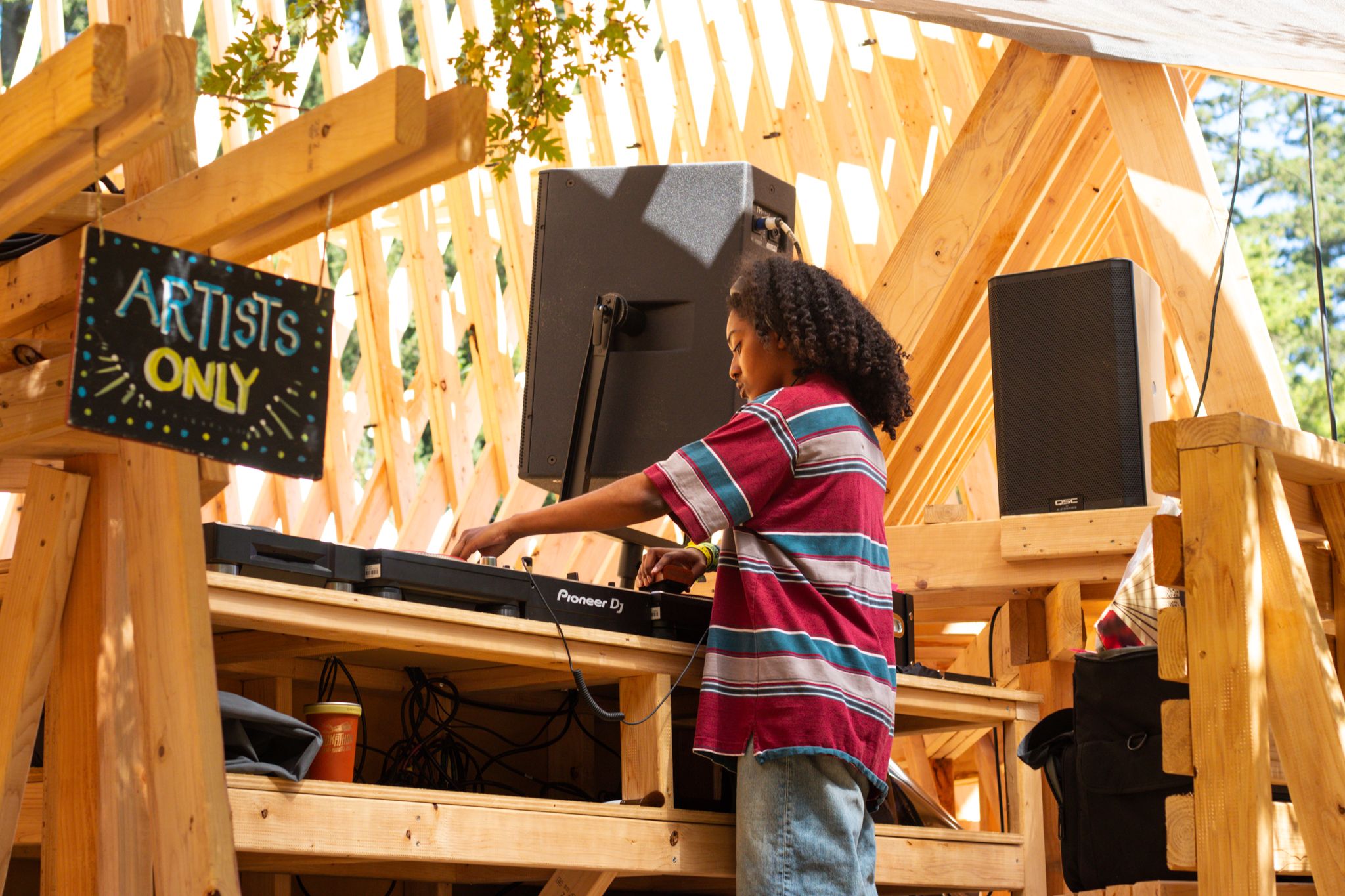 A young DJ performs at an outdoor Artists Only booth with Pioneer equipment and studio monitors under a wooden structure.