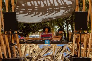 DJ performing at an outdoor festival stage with golden metallic decor, speakers, and lush green trees in the background.