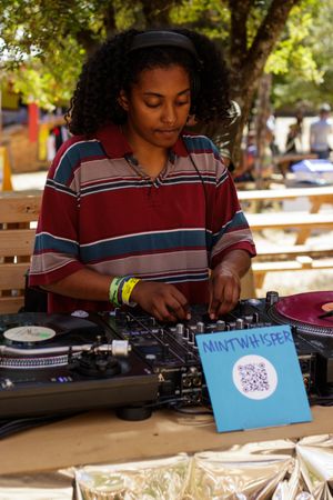 Young female DJ mixing tracks outdoors at a turntable setup with a Mintwhisper sign at a sunny event.