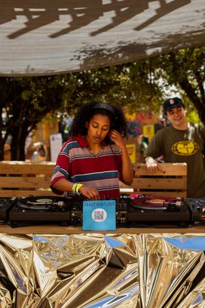 A young female DJ mixing records on turntables at an outdoor music festival with a smiling man watching in the background.