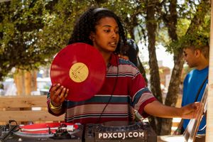 A female DJ holds a red vinyl record at an outdoor event with turntables labeled PDXCDJ.com visible.
