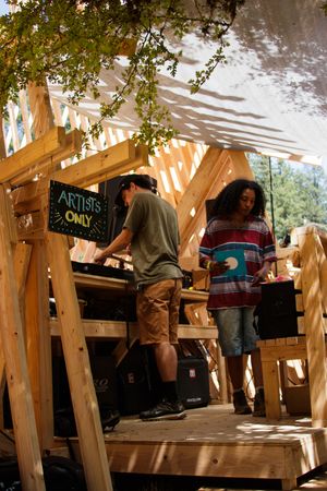 Two artists stand in a wooden structure at an outdoor festival, next to an Artists Only sign.