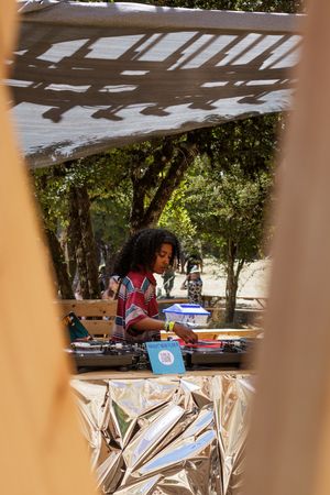 A DJ with curly hair mixing records outdoors under a pergola, surrounded by trees and colorful equipment.