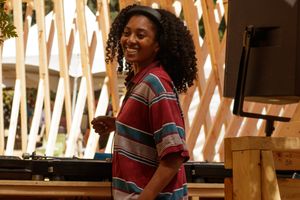 Young woman with curly hair smiling in a colorful striped shirt, standing near wooden decor outdoors.