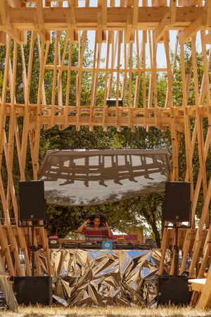DJ performing under a wooden geometric structure with large screen and metallic DJ booth at an outdoor festival.
