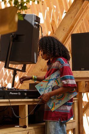 A DJ in a striped shirt holds a vinyl record while operating a Pioneer DJ mixer on a wooden stage.