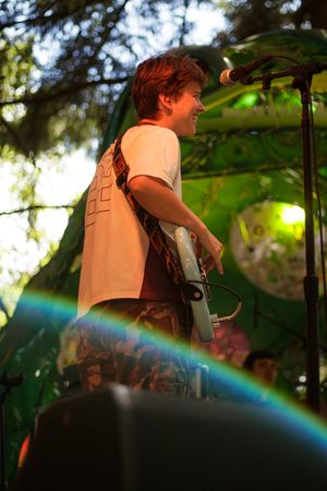 Smiling guitarist performing on outdoor stage with green backdrop, rainbow lens flare, wearing white tee and camo pants.