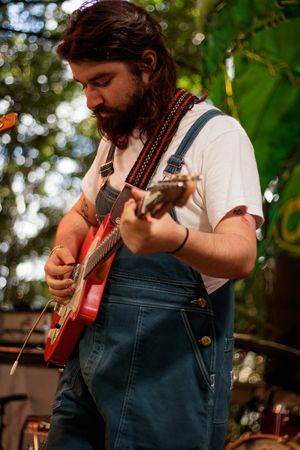 Bearded musician playing red electric guitar wearing denim overalls and white t-shirt performing outdoors at music festival.