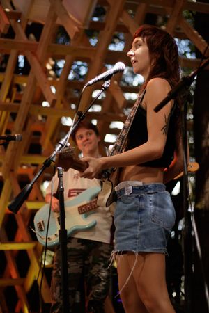 Young woman playing electric guitar and singing into microphone on outdoor stage with wooden lattice backdrop.